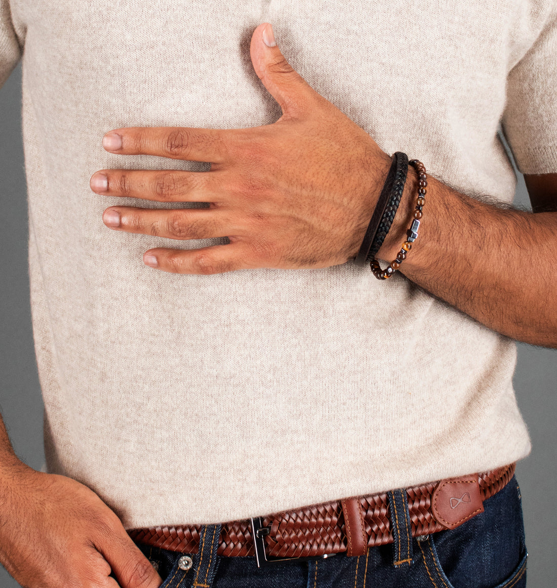 A close-up of a man wearing a light beige knit sweater, blue jeans, and a brown braided leather belt, with his right hand resting on his chest. He is wearing two black bracelets on his wrist, one of which has small gold-colored beads.