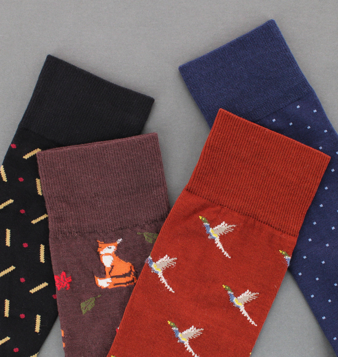 Four pairs of patterned dress socks arranged on a gray background: black with gold and red motifs, dark brown with an orange fox, rust red with white birds, and navy with small light blue dots.